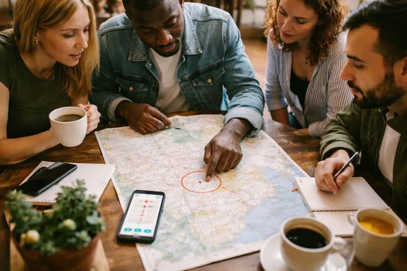 Group of people gathered around a table with a map and notebooks, pointing at a central location while discussing plans